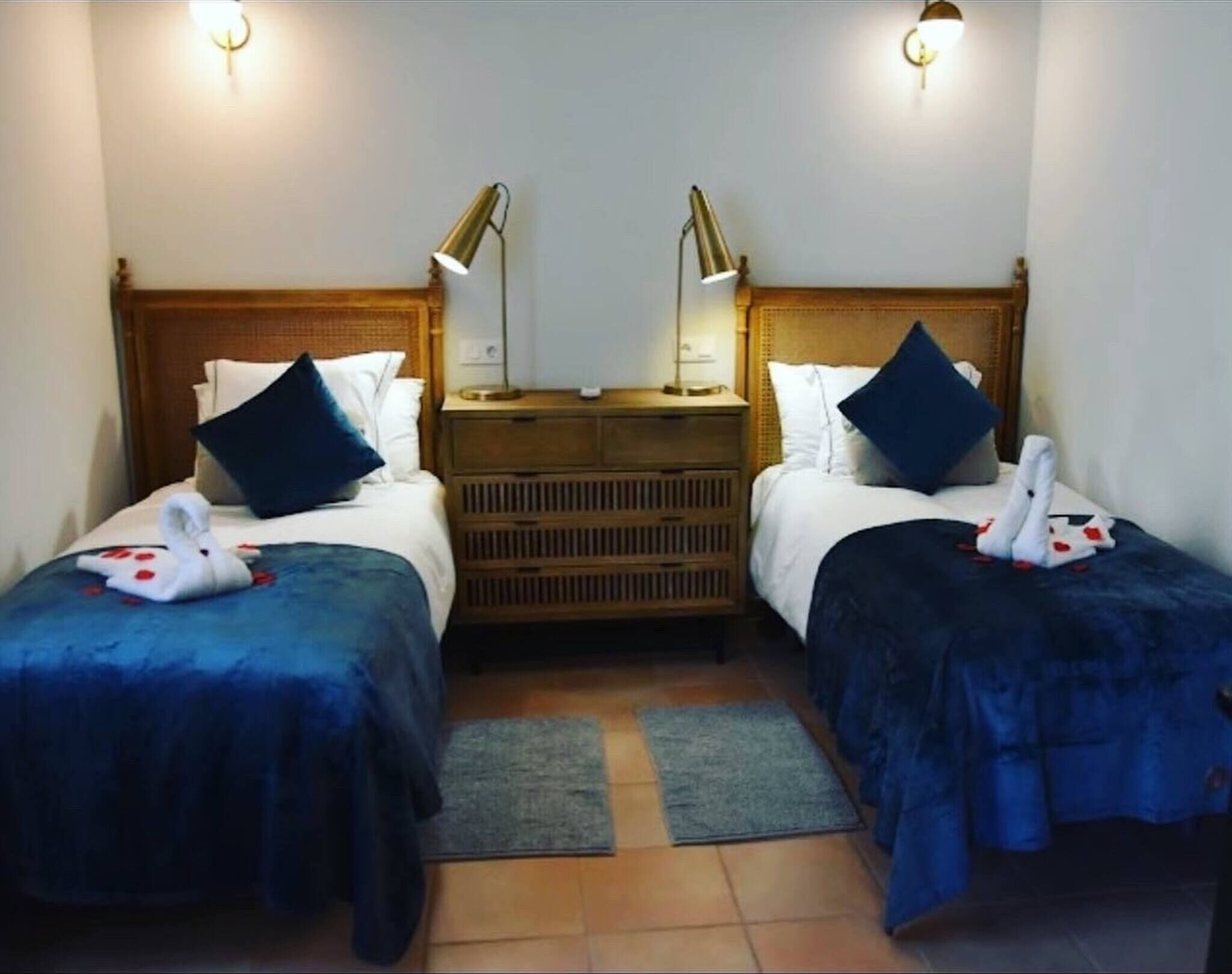 A hotel room with two beds, each with a headboard, a nightstand, a lamp, and a towel with a swan design.