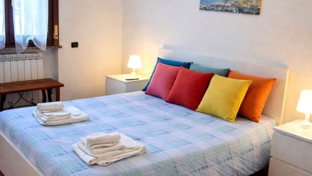 A neatly made bed with a checkered bedspread, multiple pillows in various colors, a bedside table with a lamp, and a small table with folded towels. - Brembate Comfort Home (Brembate)