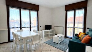 A modern living room with a dining table, chairs, a TV, a sofa, and large windows with a city view. - Brembate Comfort Home (Brembate)