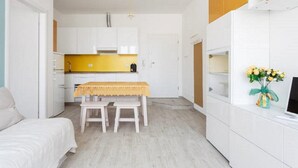 Une pièce de taille modeste comprenant un lit, une petite table à manger avec deux chaises, un coin cuisine avec des meubles blancs et un téléviseur posé sur un meuble blanc.