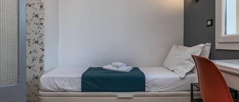 A single bed with a mattress, a teal blanket, and two pillows in a room with a wooden floor.