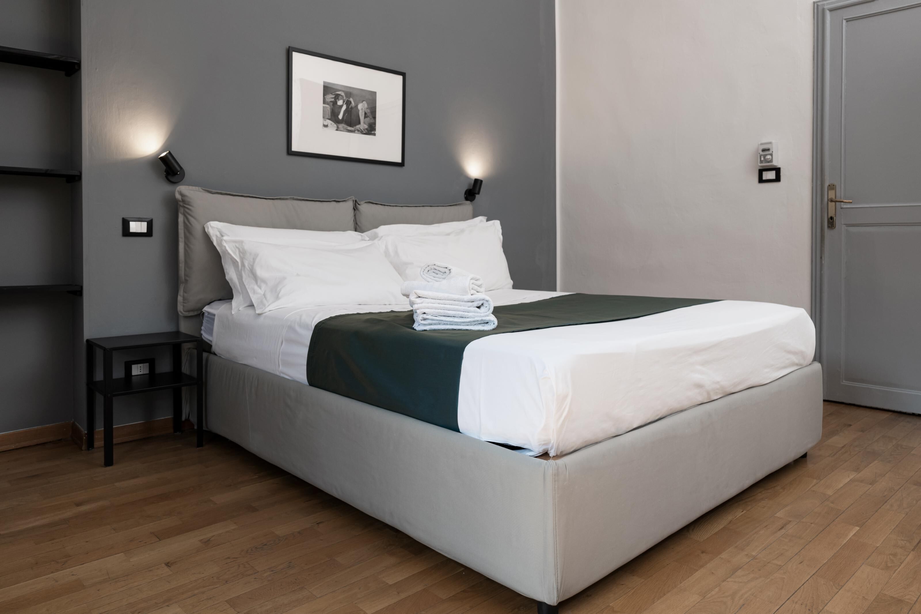 Un lit bien fait, avec du linge de lit blanc, un plaid vert foncé et une serviette pliée posée sur le côté. On y trouve une petite table de chevet noire avec une lampe, un tableau encadré accroché au mur et une porte sur la droite.