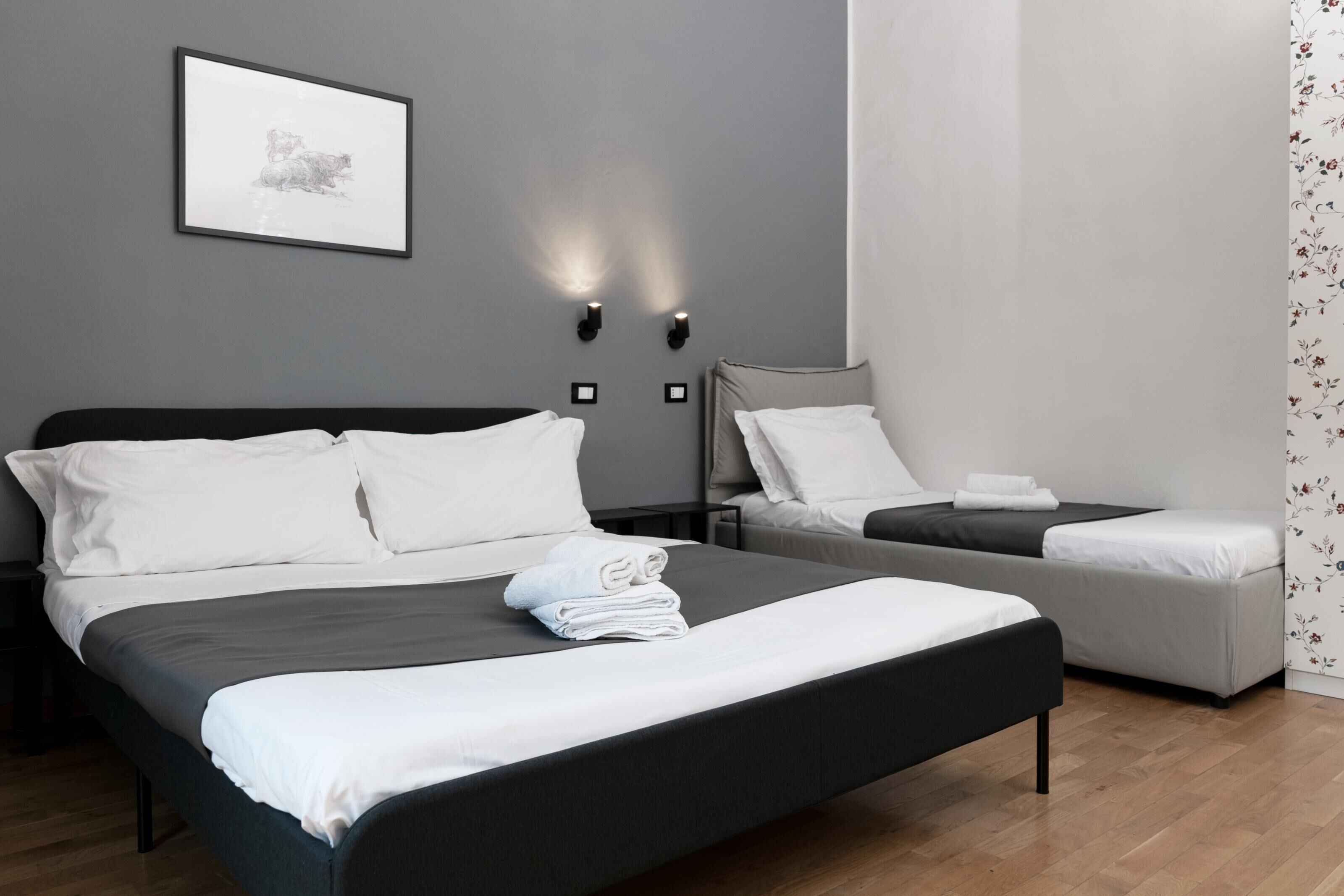 Une chambre d’hôtel moderne avec un grand lit, un plus petit lit, deux tables de chevet, une lampe fixée au mur et un tableau encadré.
