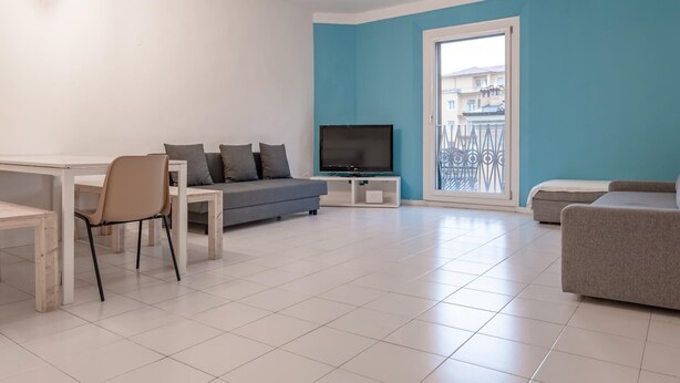 Een moderne woonkamer met een grijze bank, een kleine televisie en een eethoek met een tafel en stoelen.