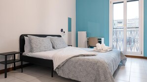 Een slaapkamer met een bed, een nachtkastje, een stoel en een raam met uitzicht op gebouwen.