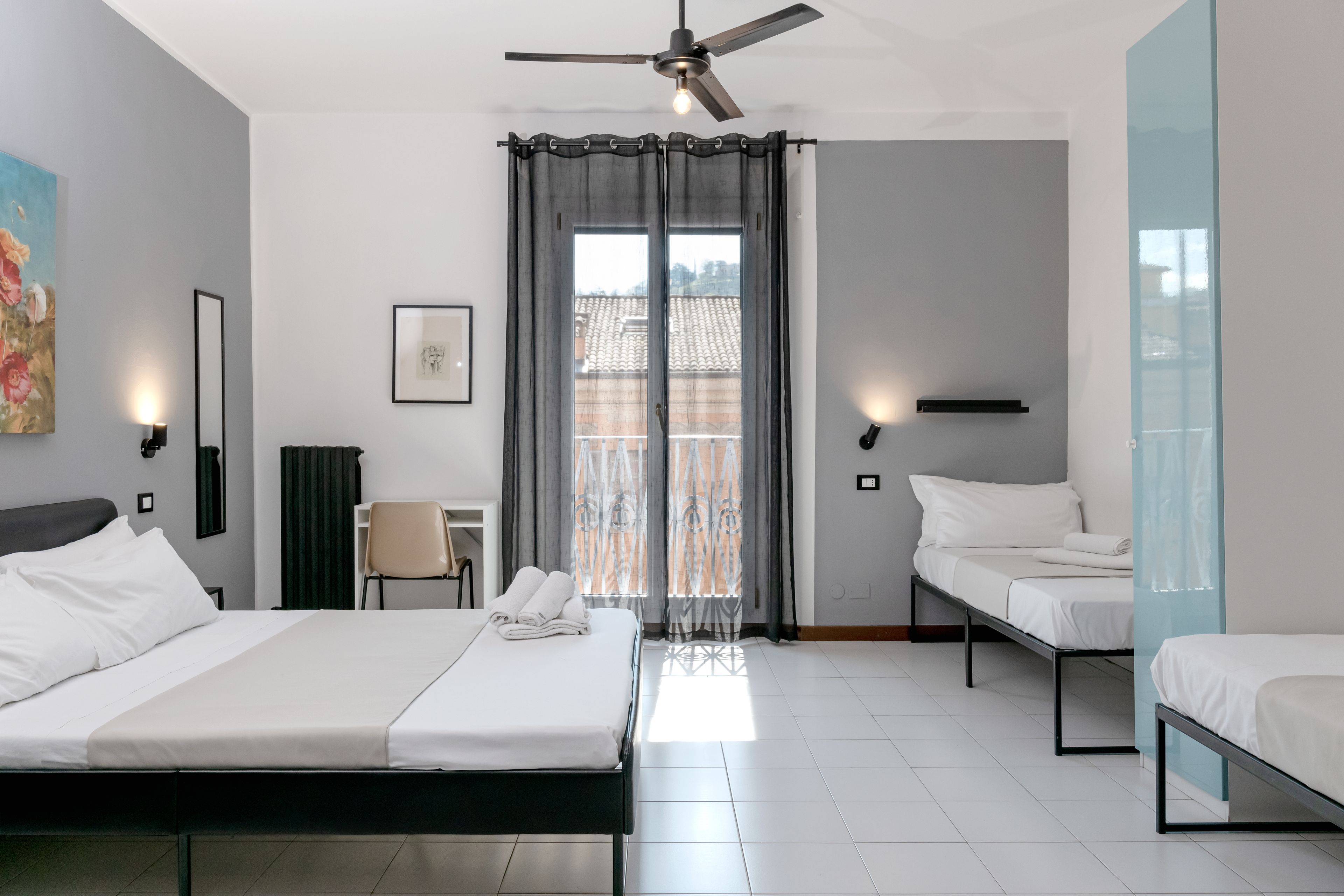 Quarto de hotel com uma cama, duas camas menores, uma cadeira, uma escrivaninha, um ventilador de teto e uma janela com cortinas.