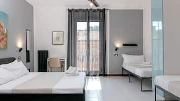 Quarto de hotel com uma cama, duas camas menores, uma cadeira, uma escrivaninha, um ventilador de teto e uma janela com cortinas.