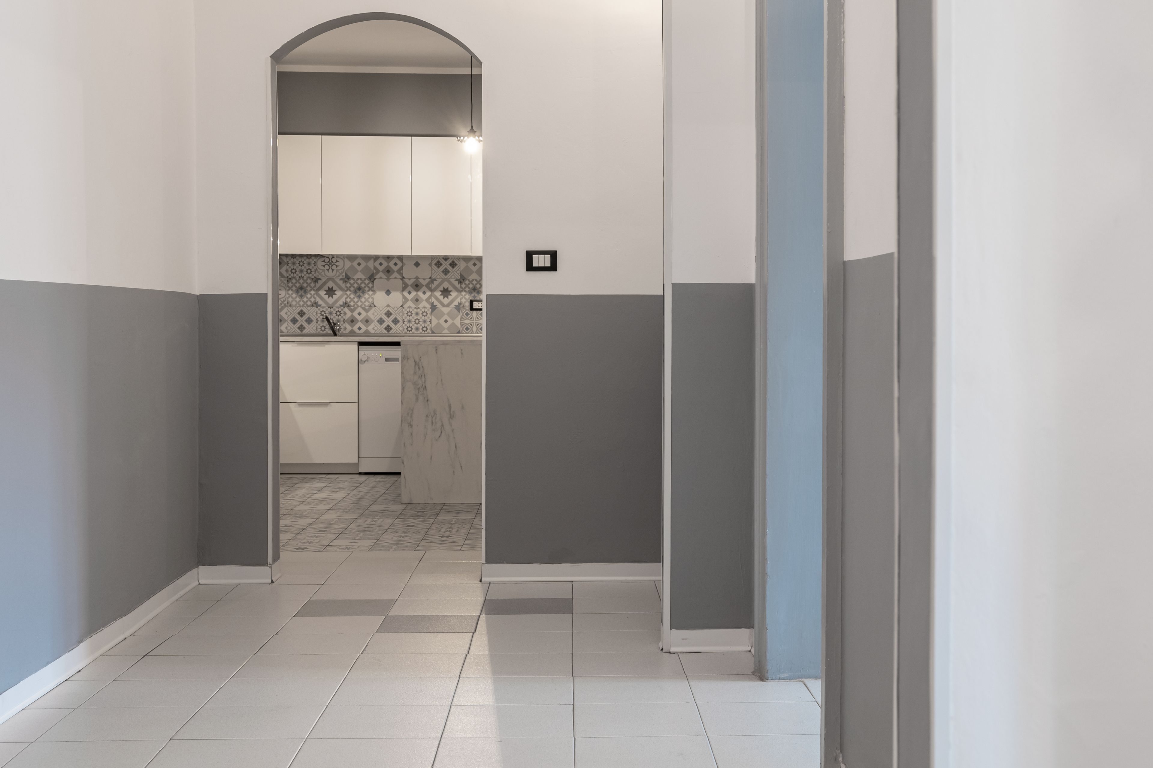 Un couloir mène à une cuisine dotée de meubles blancs, d’un pare-feu en carrelage et d’un plan de travail en marbre.