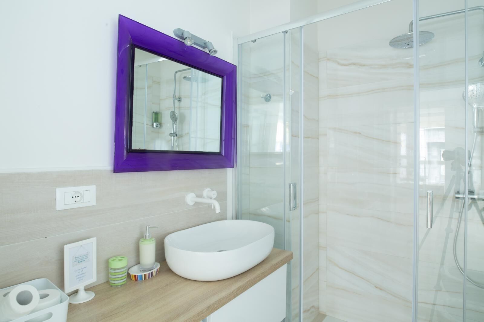 A modern bathroom with a glass shower enclosure, a white sink, and a purple-framed mirror.