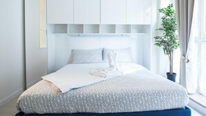 A modern bedroom with a bed, white curtains, a potted plant, and a window.