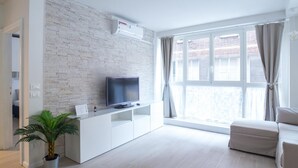 A modern living room with a flat-screen TV on a white entertainment unit, a large window with curtains, and a potted plant.