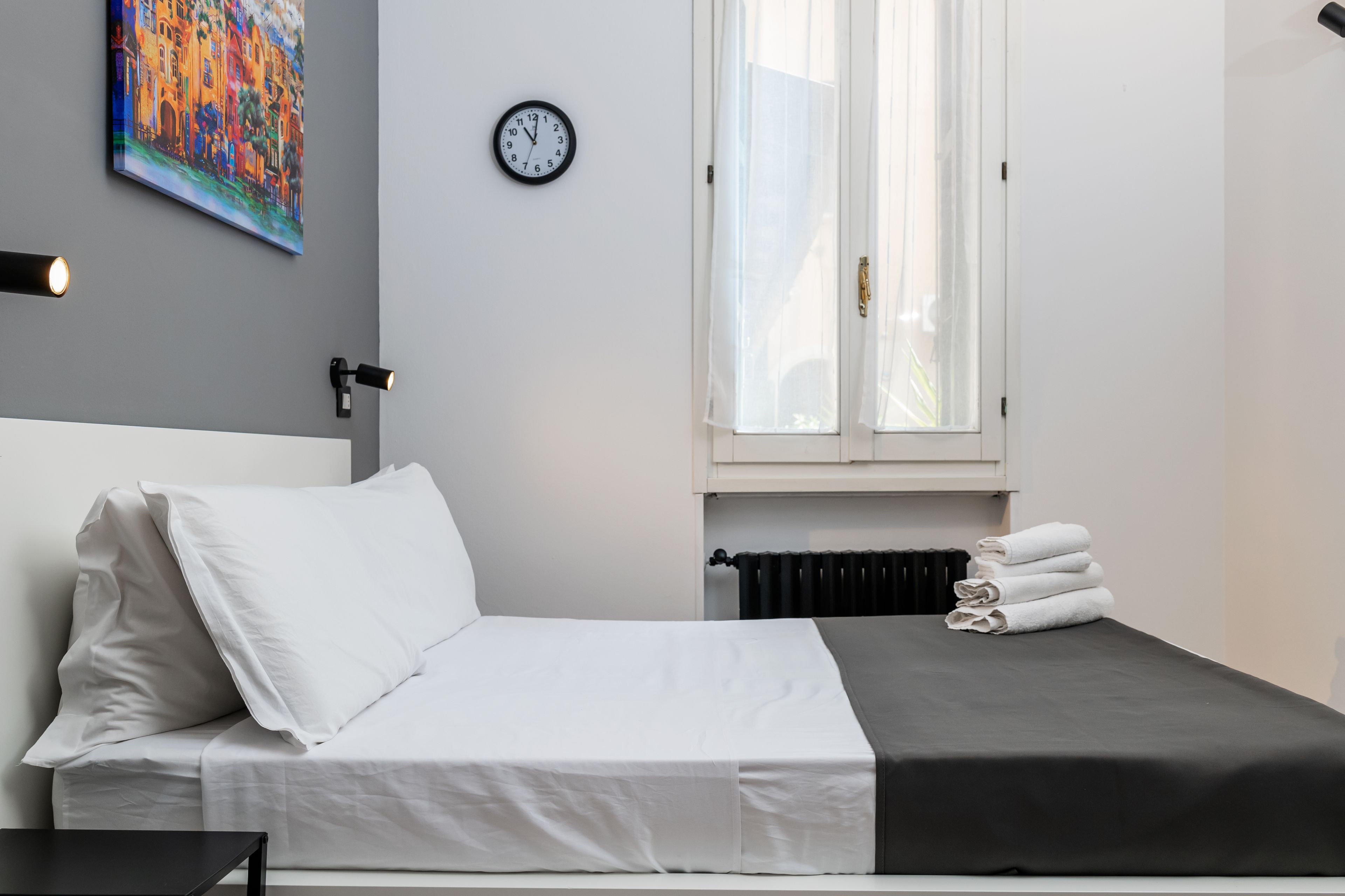 A neatly made bed with white linens, a black headboard, and a grey blanket. There is a wall clock and a painting above the bed.