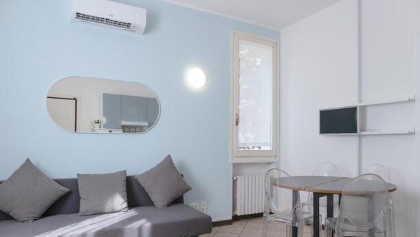 Een moderne woonkamer met een grijze bank, een ronde spiegel, een eettafel met transparante stoelen en een tv aan de muur.