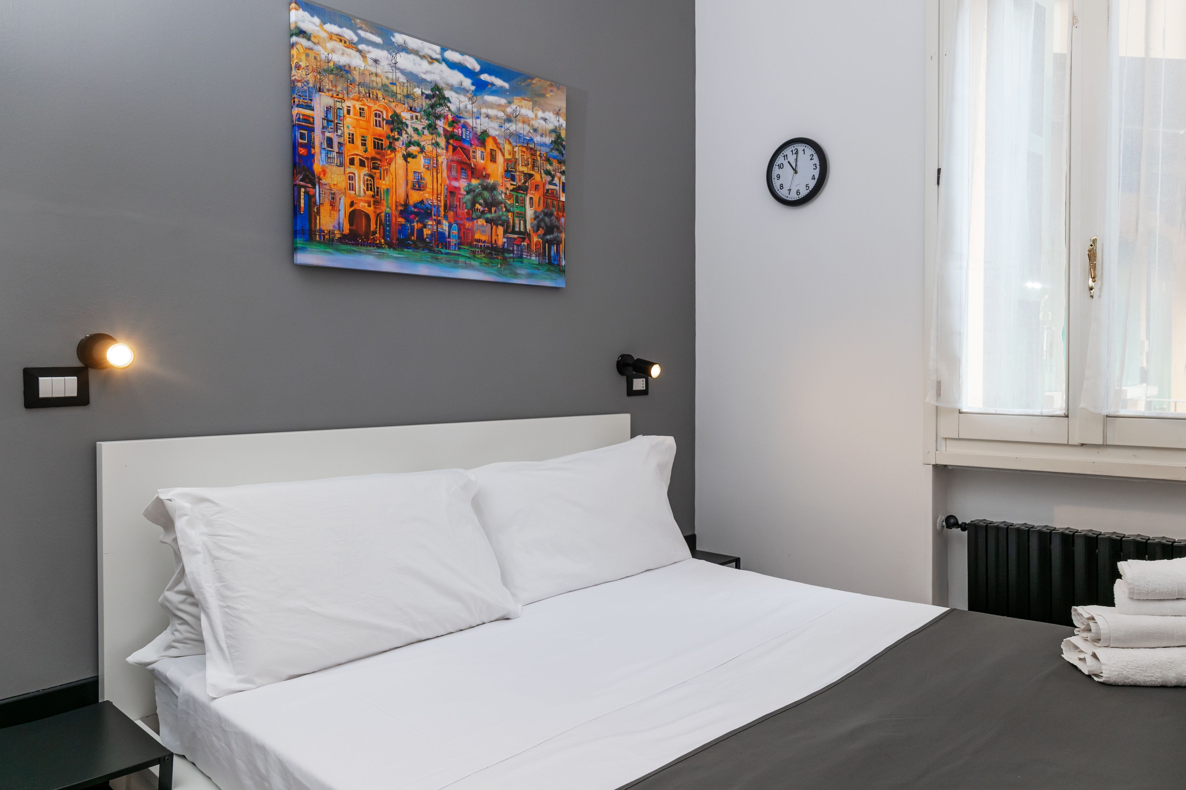 A hotel room with a bed, a painting on the wall, a clock, and a window with curtains.