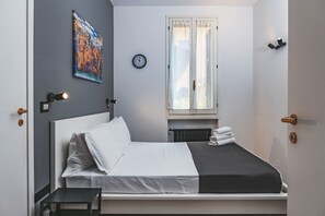 Una cama bien hecha con ropa de cama blanca y negra, una mesita de noche, un reloj de pared, una ventana con cortinas y un cuadro en la pared.