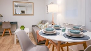 Ein moderner Essbereich mit einem runden Tisch, gedeckt für vier Personen, umgeben von grauen Stühlen. An der Wand steht eine Couch mit einer Tagesdecke und Kissen, und ein Lampenschirm auf einem Beistelltisch.