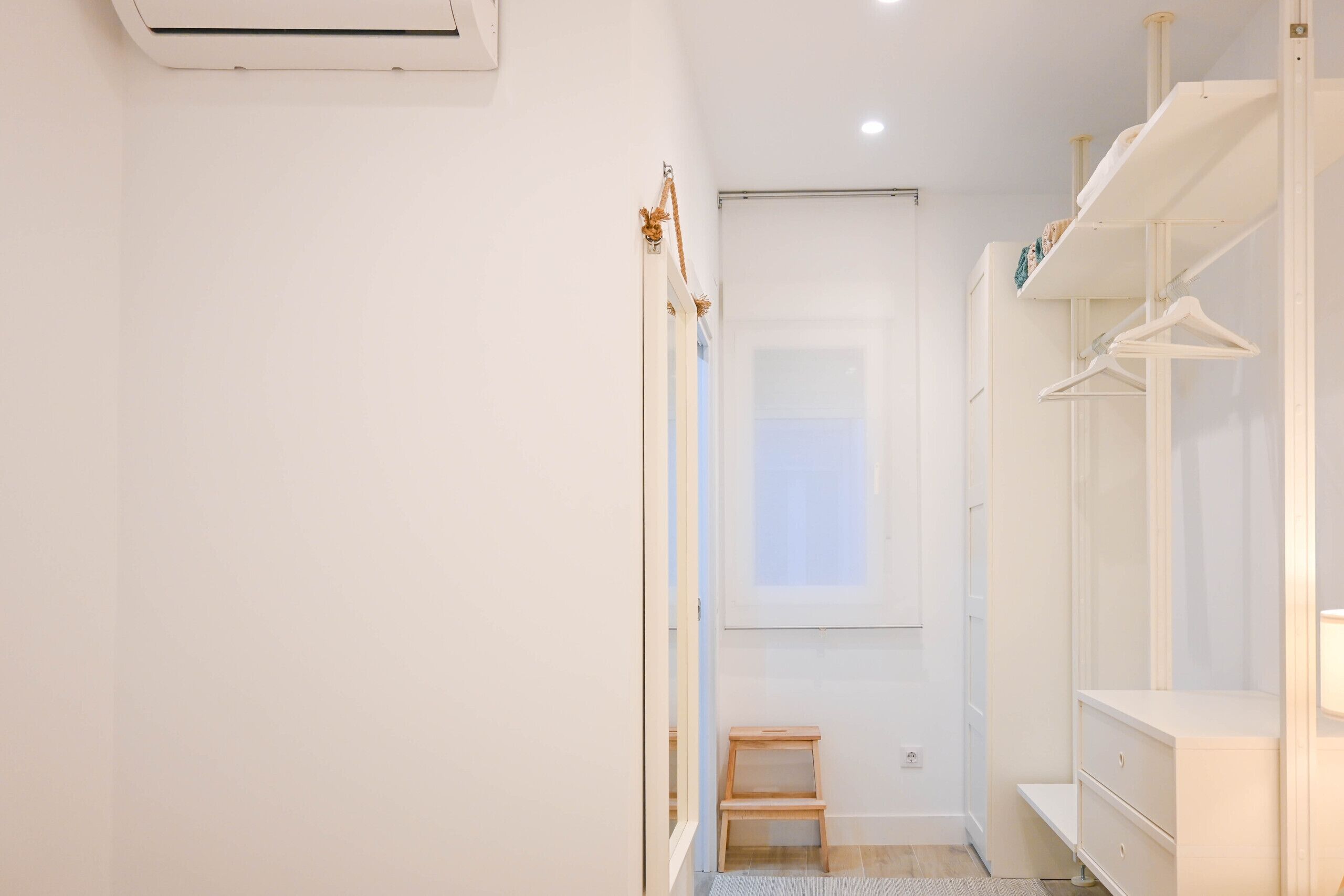 A narrow hallway with a small wooden stool, a white door, and a closet with hanging clothes.