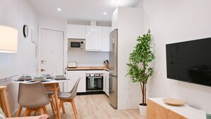 Eine moderne Küche mit weißen Schränken, einer Mikrowelle, einem Backofen und einem Kühlschrank. Ein Esstisch mit Stühlen und ein Topf mit Pflanze stehen dort. Ein Flachbildfernseher ist an der Wand befestigt.