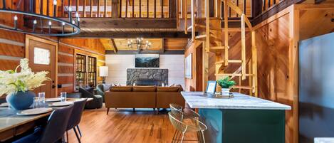 A spacious wooden cabin interior with a dining area, a living room with a sofa, a kitchen island, and a staircase leading to an upper level.