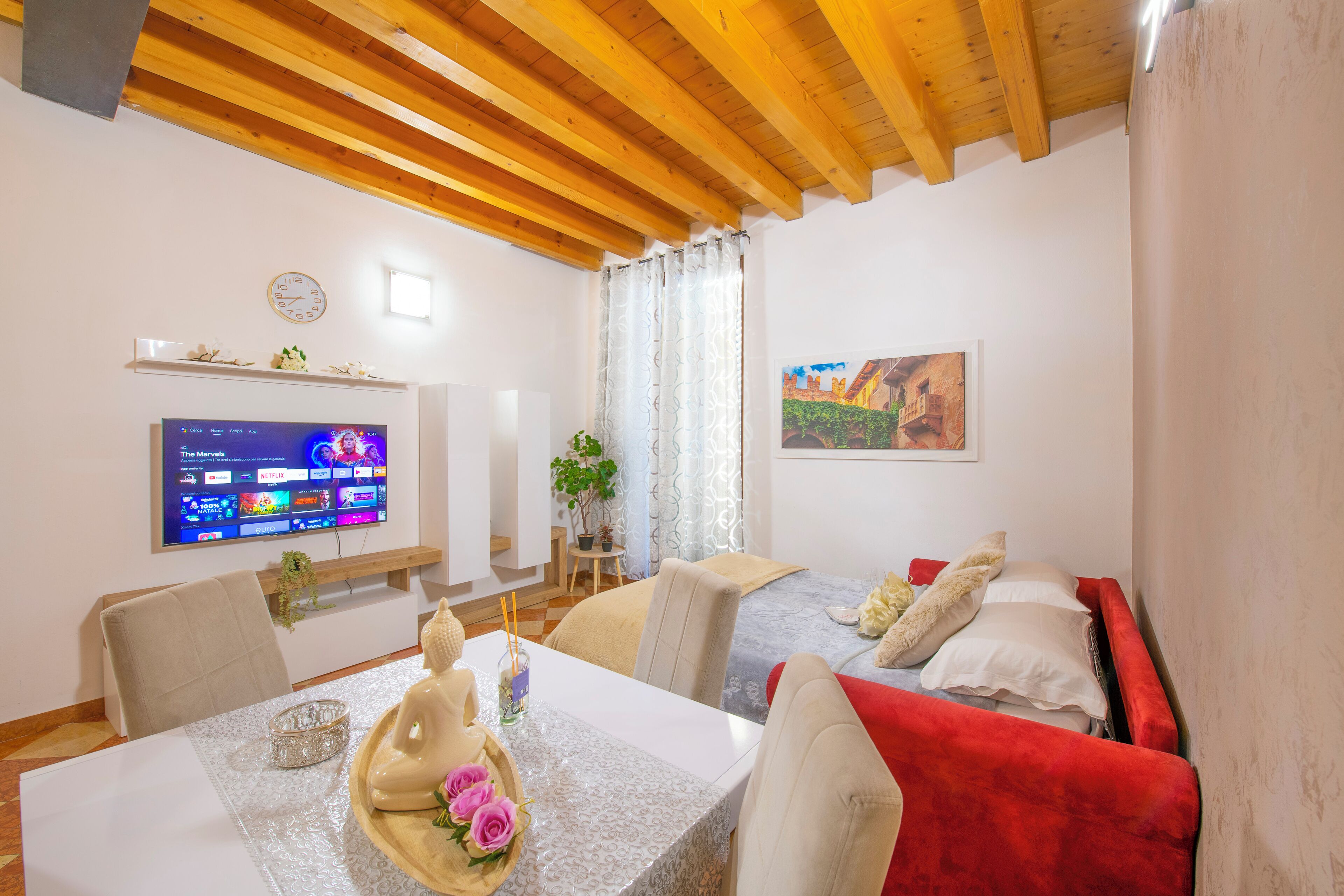 Una habitación de hotel moderna con techo de madera, televisor de pantalla plana, zona de comedor con mesa y sillas, cama con ropa de cama roja y un cuadro en la pared.