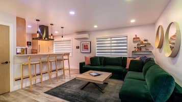 Una sala de estar moderna con un sofá verde, una mesa de centro y una zona de cocina con armarios de madera y taburetes de bar.
