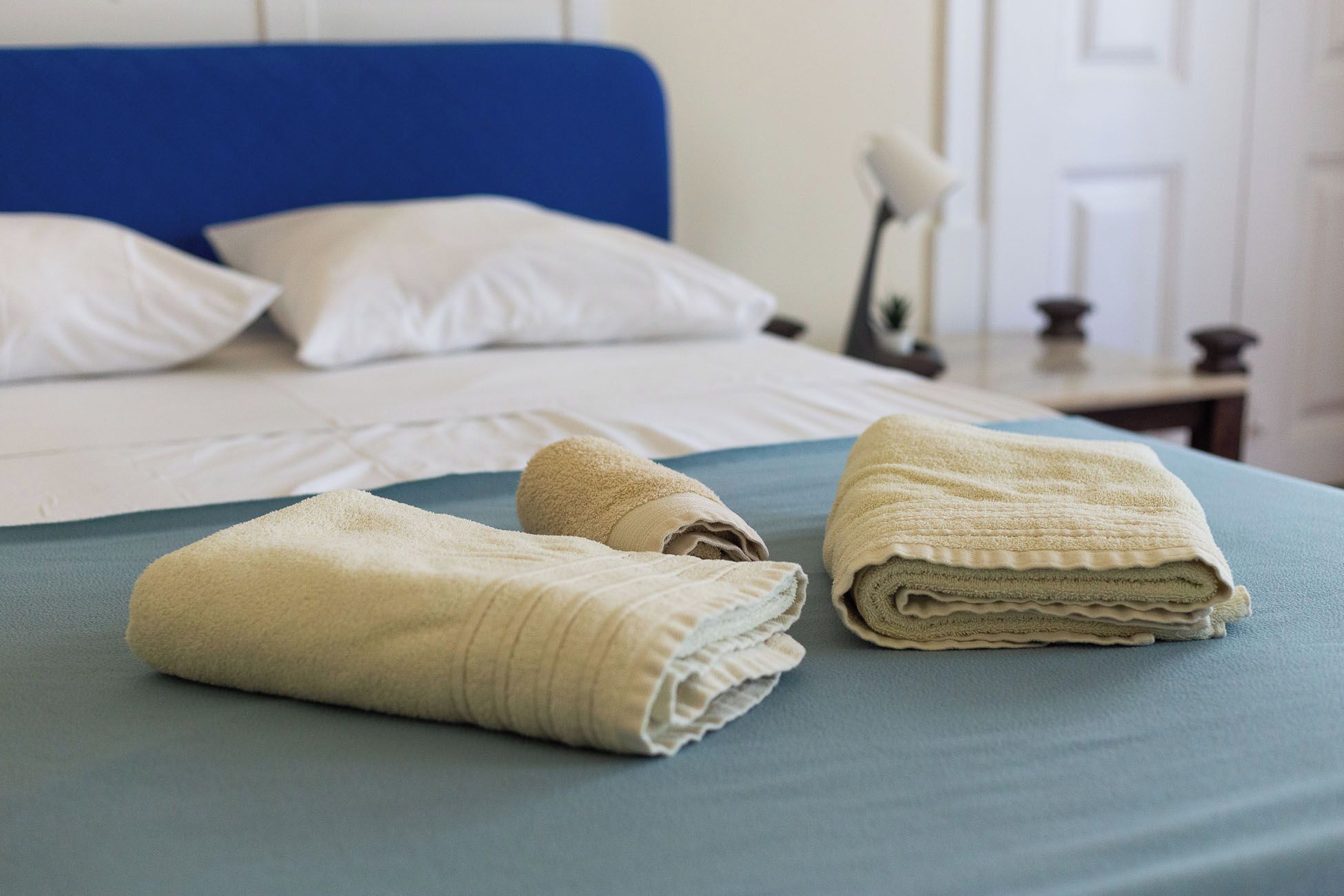 Una cama bien hecha con cabecera azul, almohadas blancas y toallas dobladas.