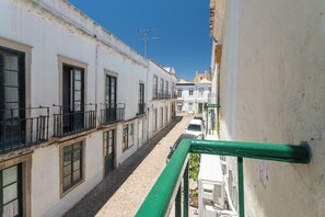 Una calle estrecha flanqueada por edificios, un coche blanco estacionado a la derecha y una barandilla verde.