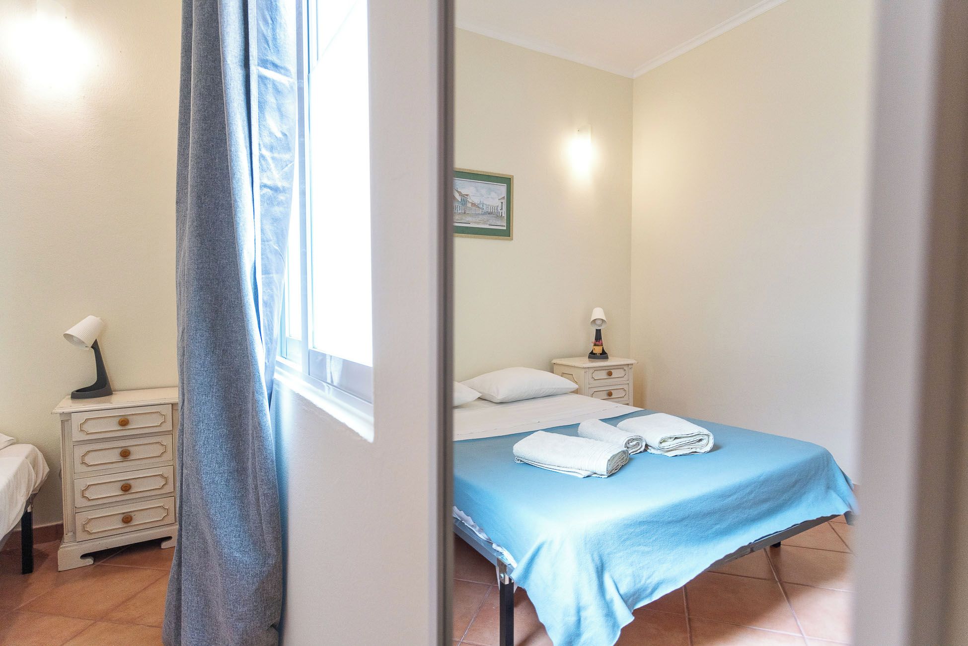 Quarto com cama, mesa de cabeceira, luminária e janela com cortinas.