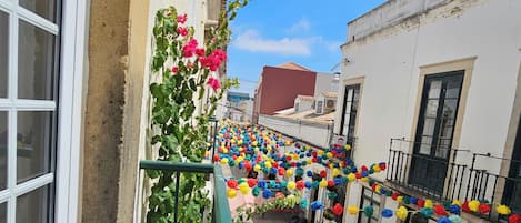 Une ruelle étroite, avec un balcon orné de fleurs en papier aux couleurs vives, un immeuble avec un toit rouge et un mur blanc percé d’une fenêtre.