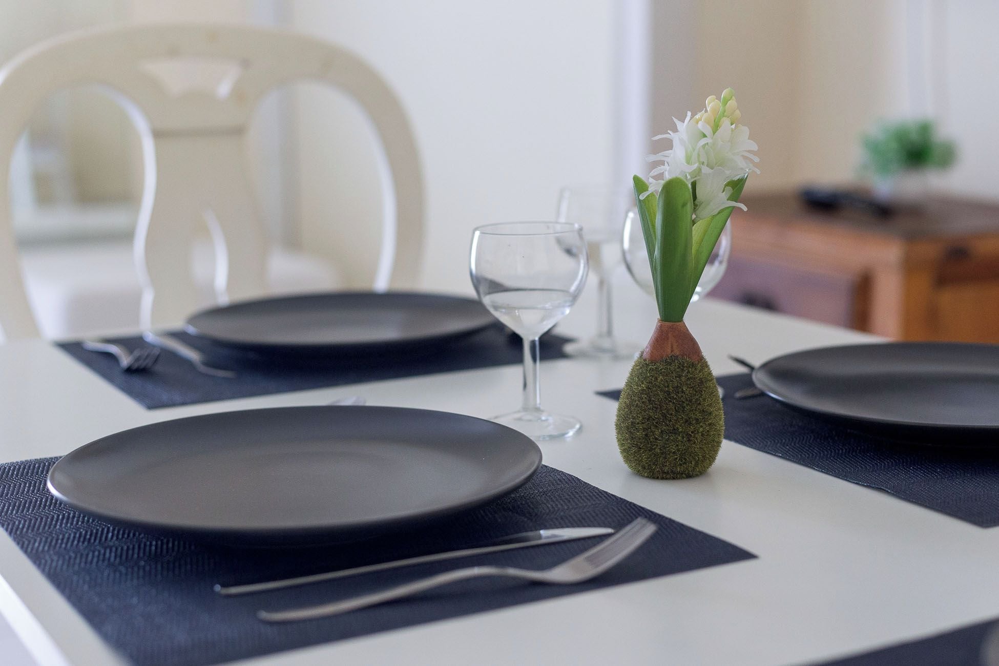 Taula de dinar posta amb plats foscos, coberts i un got. Un vas petit amb flors blanques hi és col·locat.