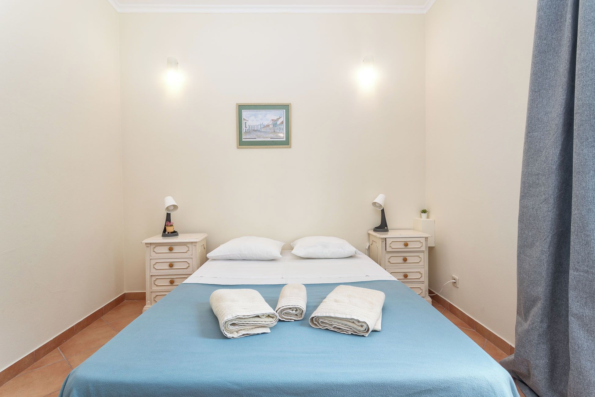 Habitación de hotel con cama, dos mesitas de noche, una lámpara de noche, un cuadro en la pared y una ventana con cortinas.