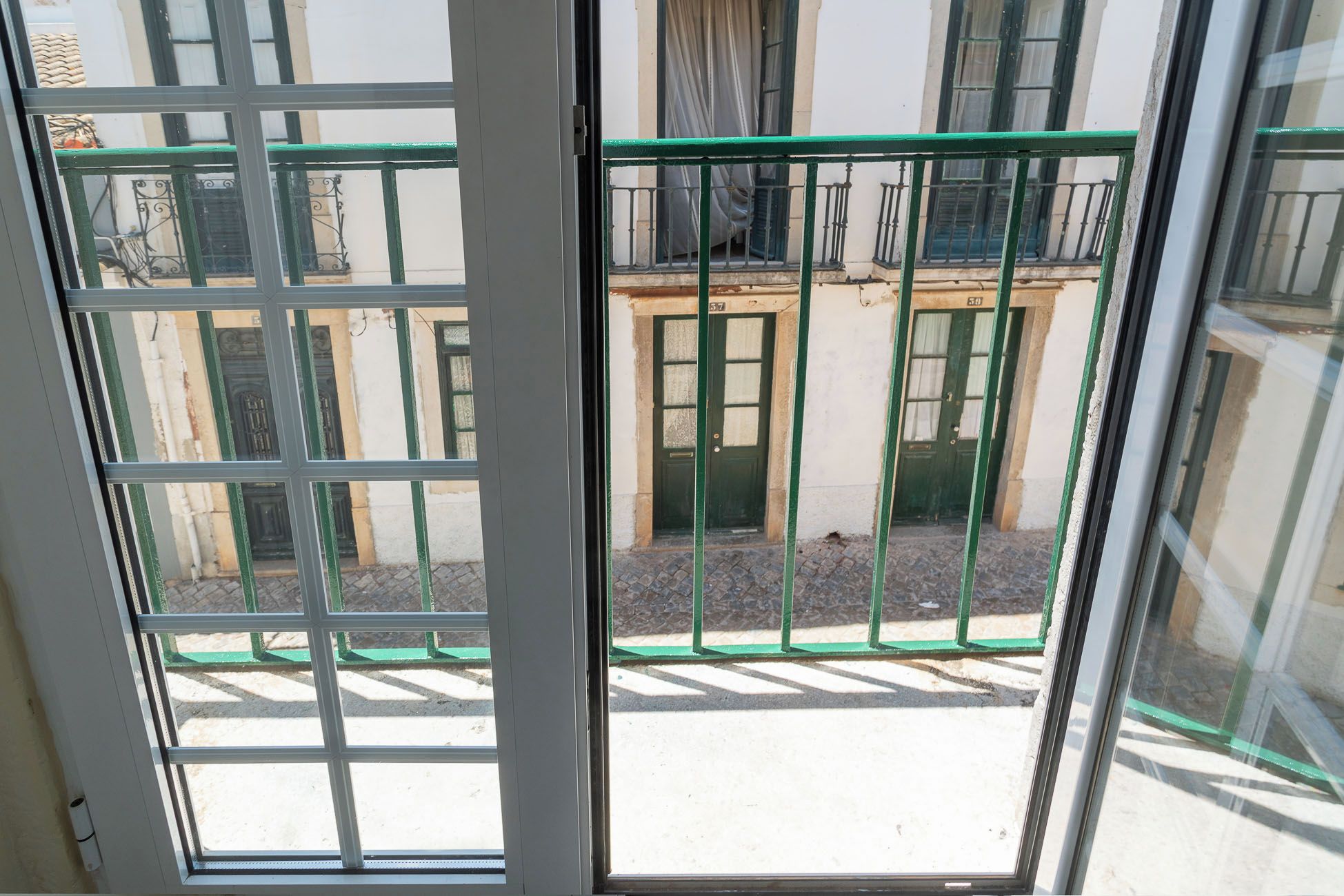 Vista attraverso una finestra con cancello in metallo, un edificio con una porta verde e terreno lastricato.