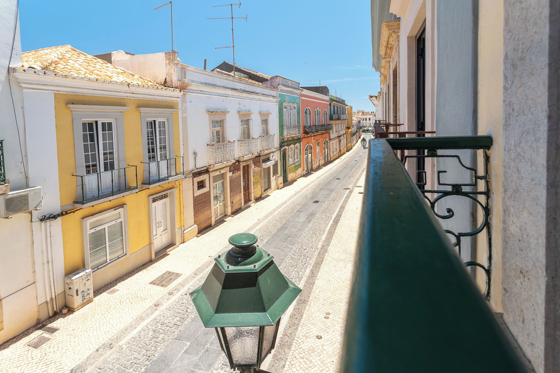 Una calle estrecha flanqueada por edificios, una baranda de metal verde y un cielo despejado.