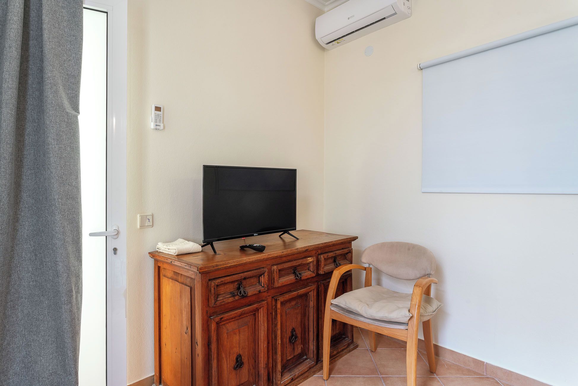Quarto com televisão sobre um armário de madeira, uma cadeira e um quadro branco na parede.