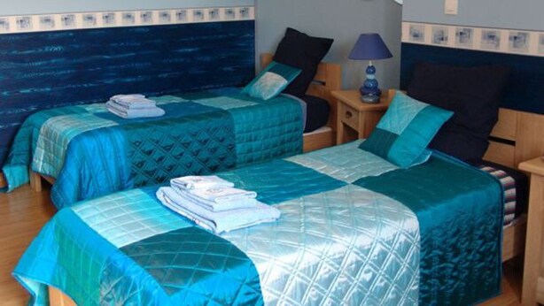 Une chambre avec deux lits, chacun agrémenté de draps et d’oreillers bleus, une petite table de chevet avec une lampe, et un mur orné d’un motif ondulé.