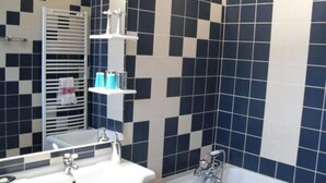 Une salle de bain avec un lavabo blanc, une baignoire et un miroir au-dessus du lavabo. On y trouve un porte-serviettes sur lequel est suspendue une serviette, ainsi qu’une étagère avec des articles de toilette.