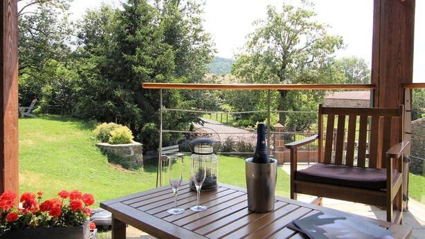 A wooden table with two wine glasses and a metal container, a wooden chair, and a view of a green landscape with trees and a stone structure. - Spacious suite with wellness, parking, wifi, in Saint-Privat-d'Allier (Saint-Privat-d'Allier)