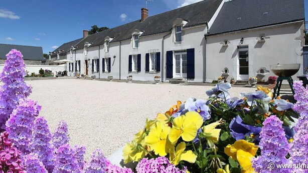 A row of white buildings with dark shutters, a gravel courtyard, and a variety of flowers in the foreground.