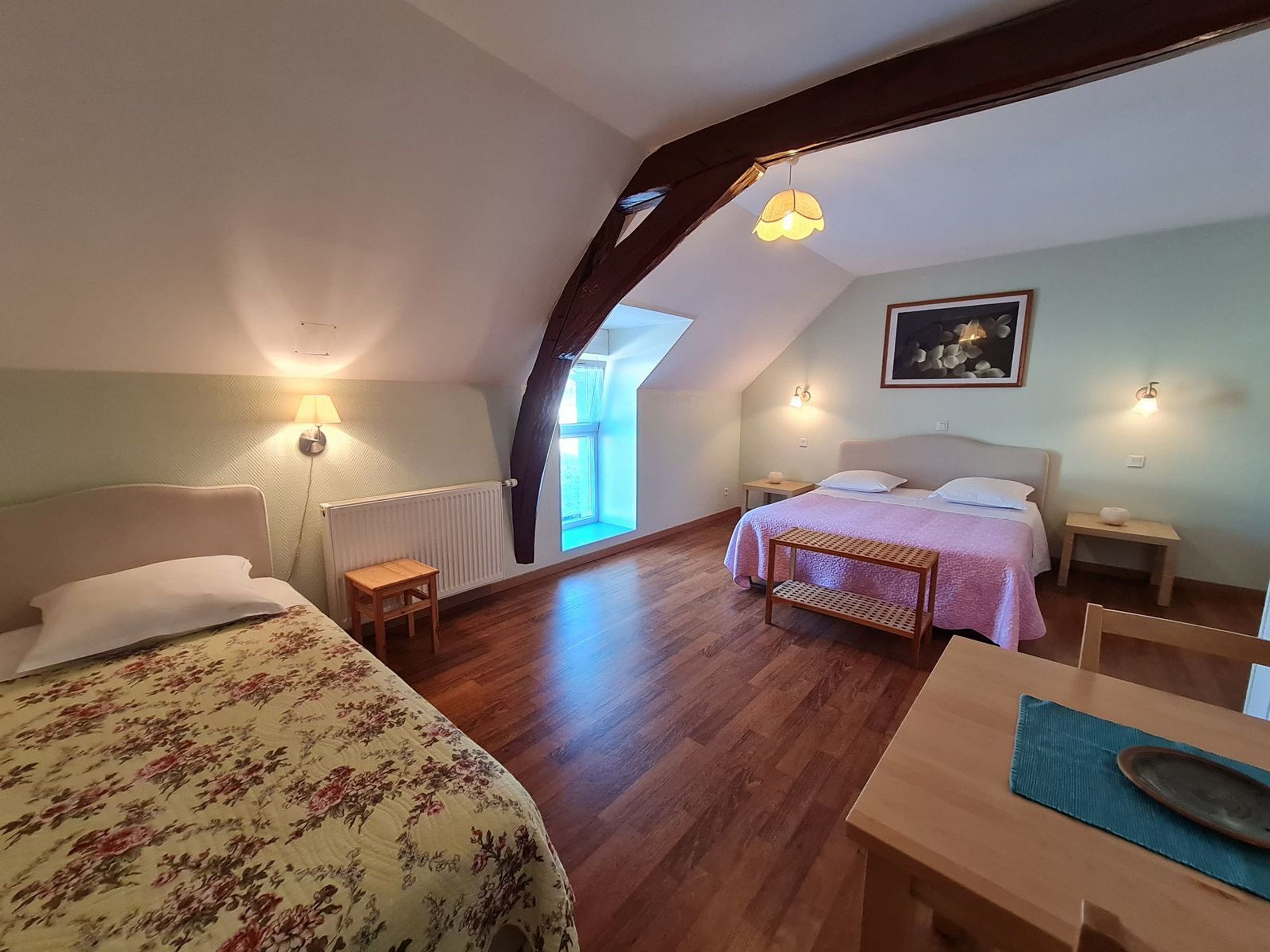 Un dormitorio con techo inclinado, una cama con colcha estampada, una mesita de noche, un taburete de madera pequeño, una mesa con un plato y una lámpara fijada a la pared.