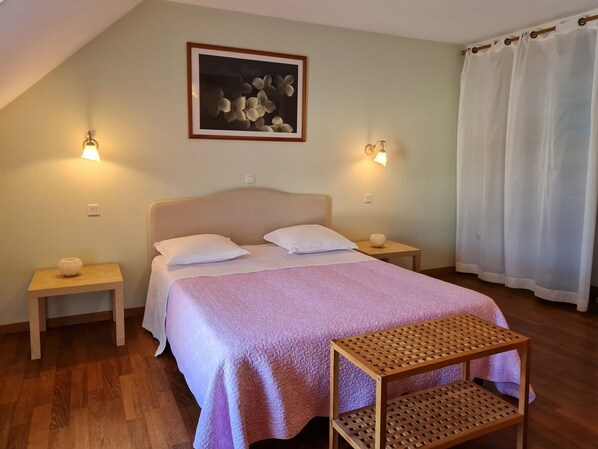 A bedroom with a bed, two bedside tables, a small wooden bench, a wall-mounted picture, and a window with white curtains.