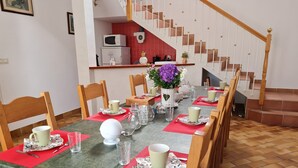 A dining area with a long table set for a meal, wooden chairs, a staircase, and a kitchen in the background.