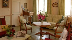 Ein gemütliches Wohnzimmer mit einer Couch, Sesseln, einem Tisch mit Blumenvase, einem Bücherregal und einem Fenster mit Vorhängen.
