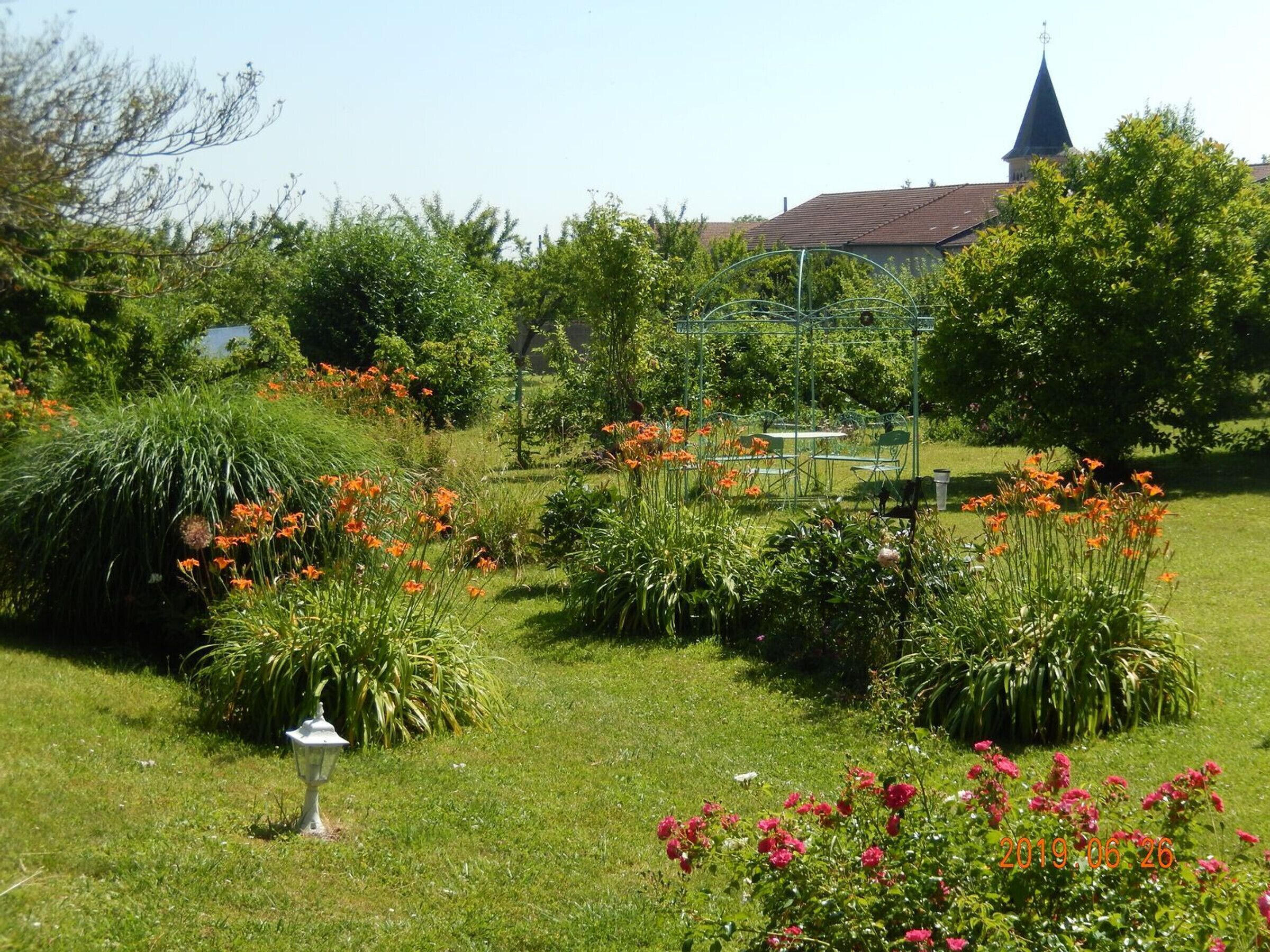 Un jardín con diversas plantas y flores, un pabellón y una iglesia al fondo.