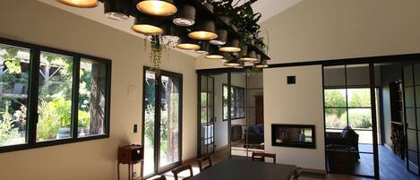 A modern dining area with a long table, multiple chairs, pendant lights, and large windows offering views of greenery.