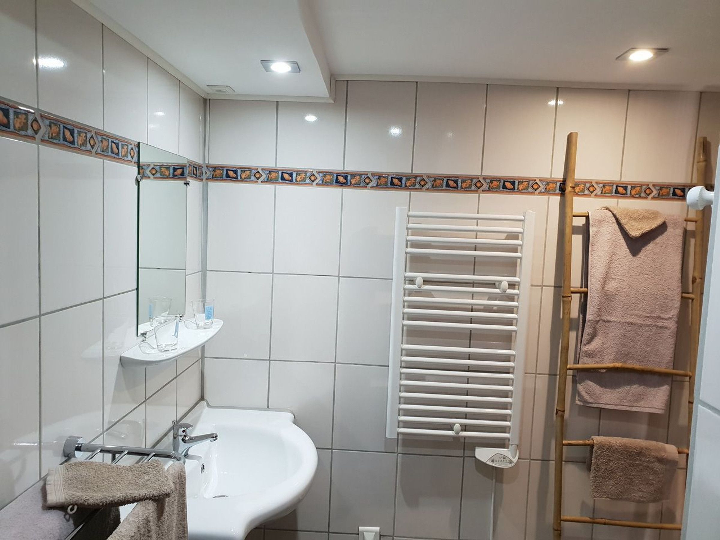 Ein Badezimmer mit weißem Waschbecken, Spiegel und gefliesten Wänden. Ein Handtuchhalter mit zwei Handtüchern und eine Holzleiter mit zwei Handtüchern.