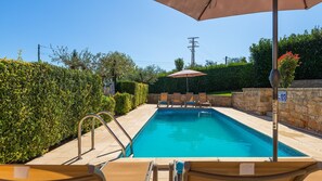 Une piscine rectangulaire dont les bords sont recouverts de carreaux, entourée d’un mur de pierre et équipée d’une échelle en métal. On y trouve des chaises longues et un grand parasol qui offre de l’ombre.