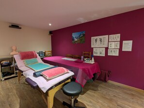 A massage therapy room with a wooden massage table, a foot massage table, and a foot massage chair. There are two wooden chairs, a shelf with massage equipment, and a wall with foot massage illustrations. - Family room with pool and SPA in Anould (Anould)