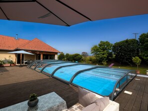 A covered swimming pool with a wooden deck, a patio table, and a house in the background. - Family room with pool and SPA in Anould (Anould)