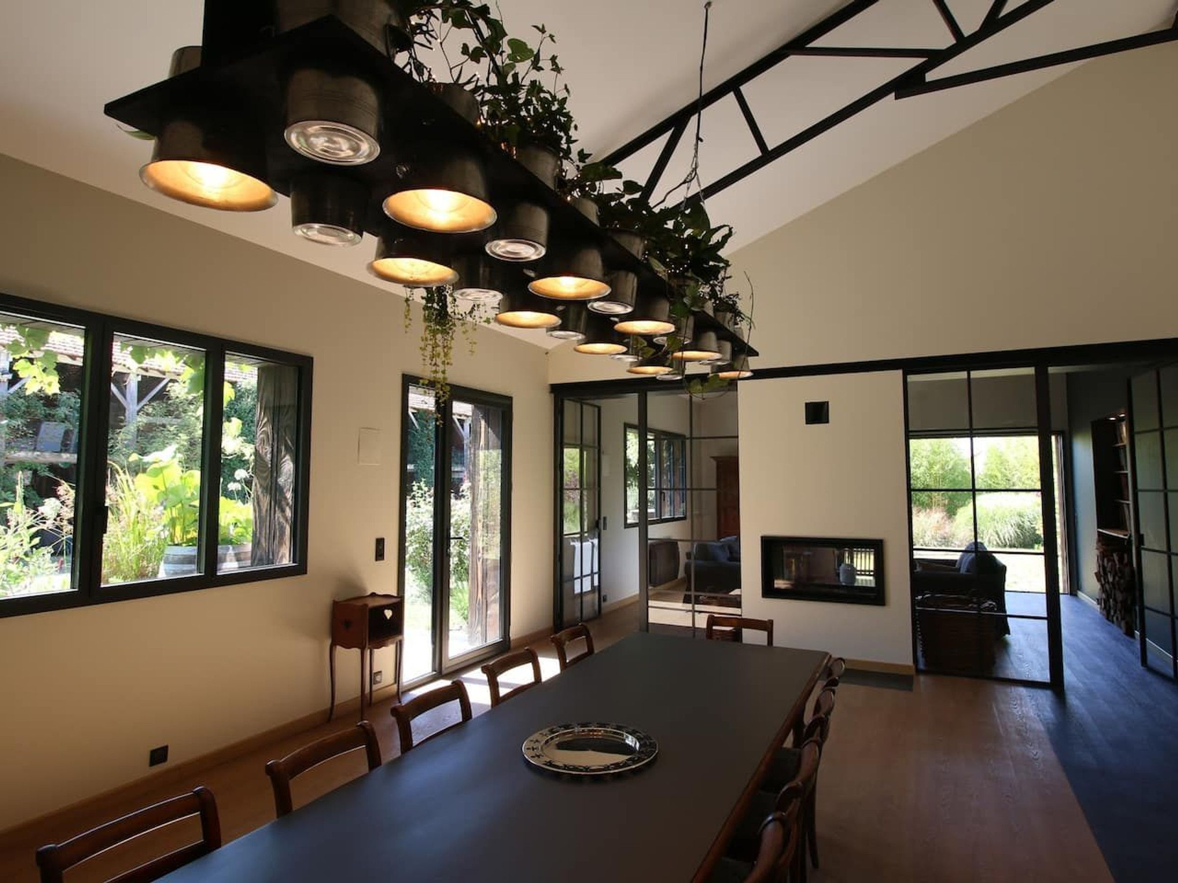 Un comedor moderno con una mesa larga, múltiples sillas, plantas colgantes y amplios ventanales con vistas a zonas verdes.