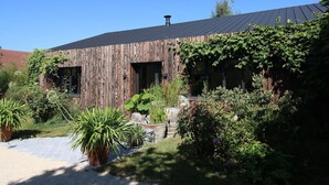 Un bâtiment de style rustique, avec une façade en bois, de grandes fenêtres et un jardin agrémenté de diverses plantes ainsi qu’un chemin pavé.